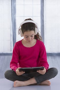 Student sitting on the floor wearing headphones and reading from a tablet computer.